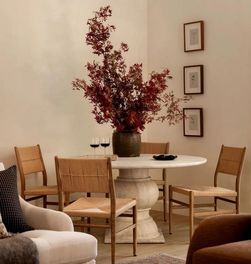 A dining corner with a round stone pedestal table, woven rush chairs and a tall vase of red maple branches