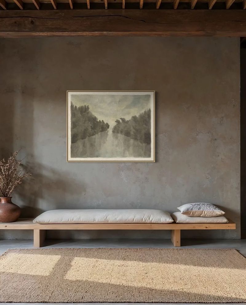 A misty river landscape painting hung above a long wooden bench in a room with plaster walls and a timber beam